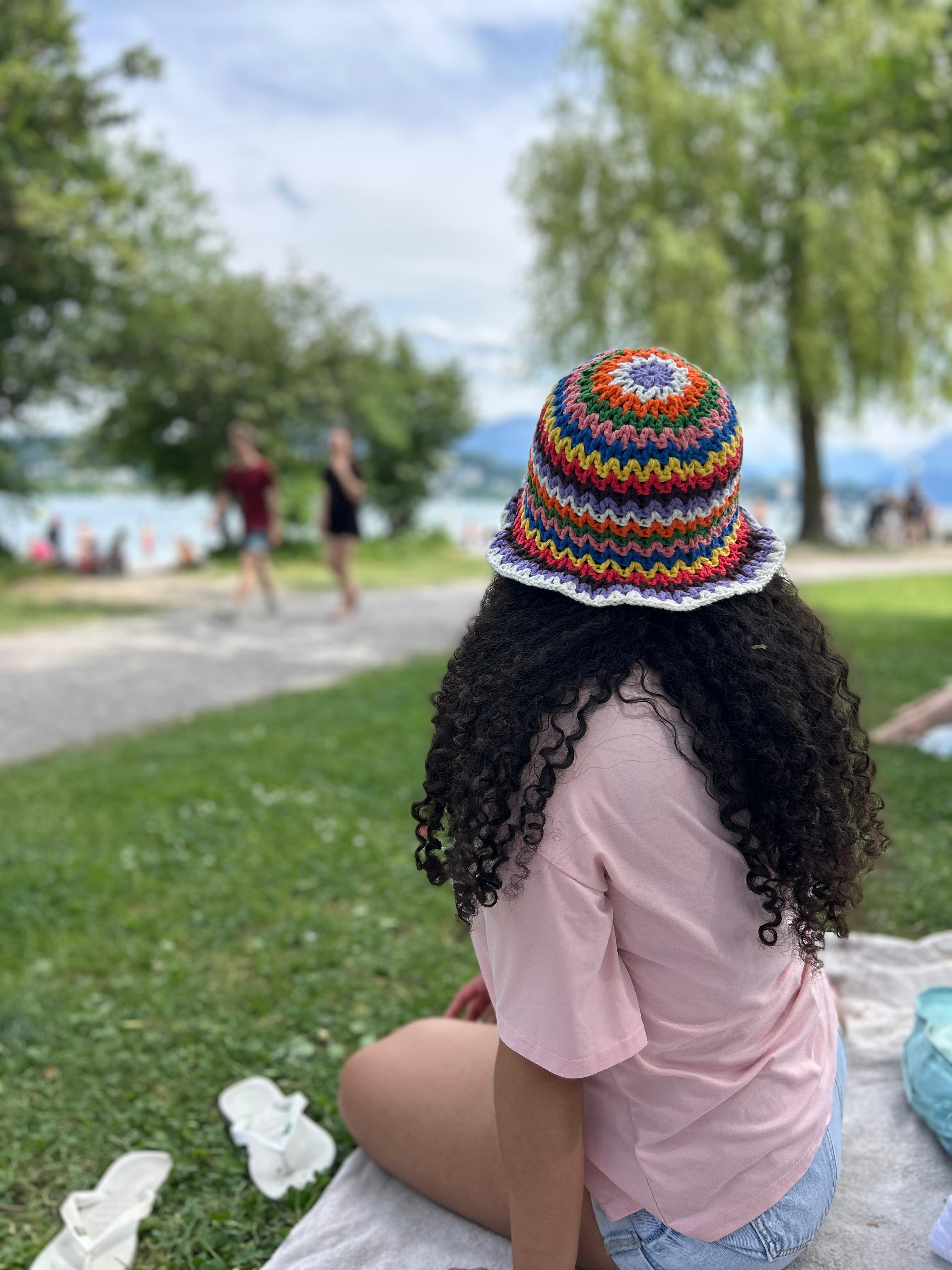 A person wearing a colorful rainbow striped OUI crochet bucket hat by a lake